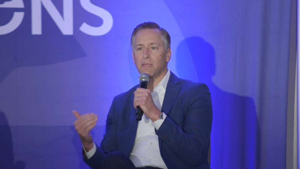 A person in a blue suit speaks into a microphone on stage with a blue backdrop.